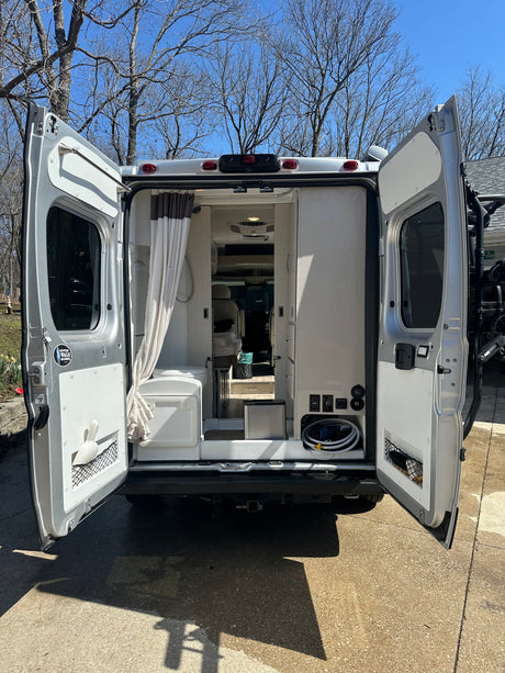 Rear view of an RV rental camper van with open doors, revealing its compact interior including a shower and utility hookups.