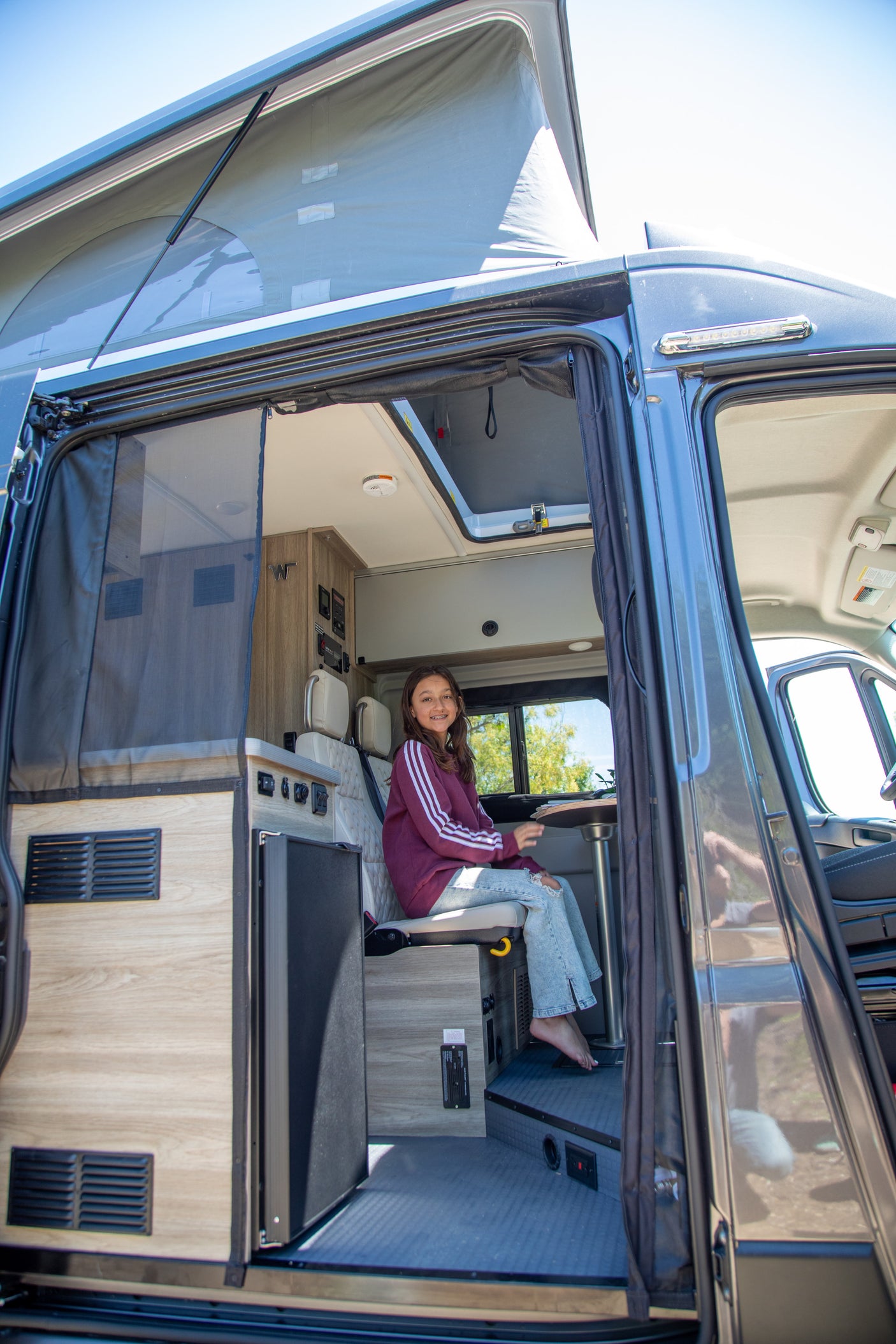 A girl smiles from inside a grey RV rental with its pop-top roof extended, showcasing the light wood interior.
