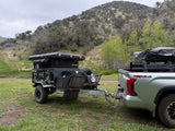 An overland RV rental trailer with rooftop tent hitched to a truck, parked in a grassy mountain area.