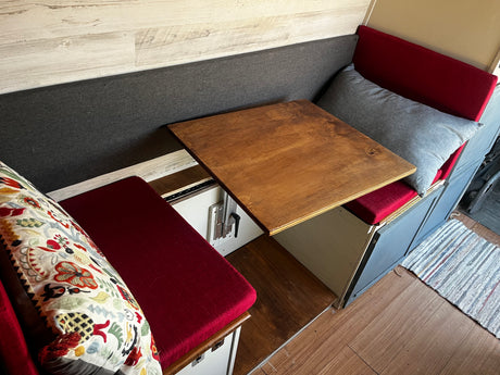 RV rental campervan dinette: a wooden table sits between two red-cushioned benches, backed by a grey fabric wall panel.