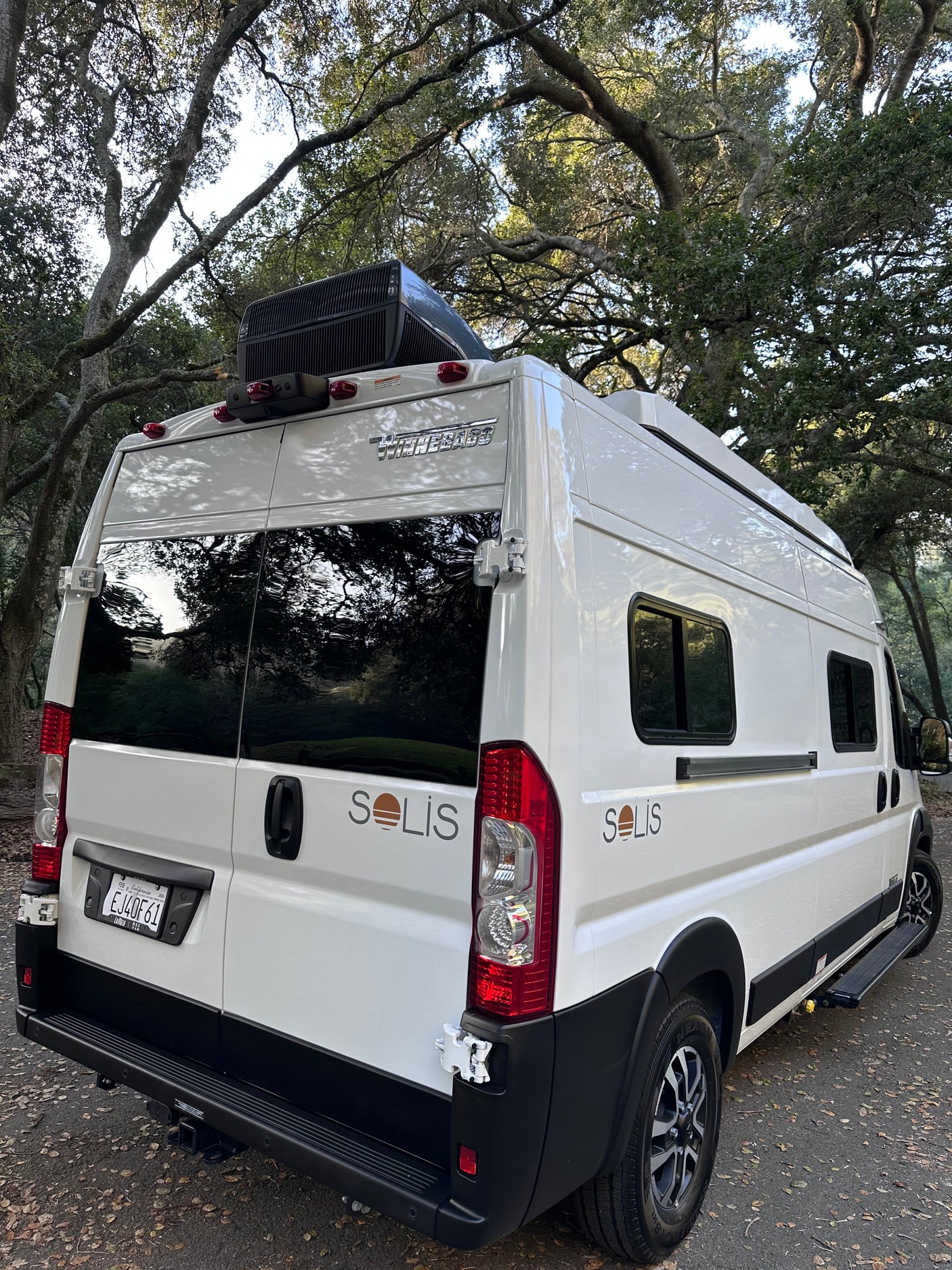 This white Winnebago Solis camper van, ideal for rv rental, is parked beneath large trees, ready for adventure.
