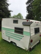 A vintage white & green travel trailer RV, ready for an rv rental, parked in a grassy yard under a cloudy sky.