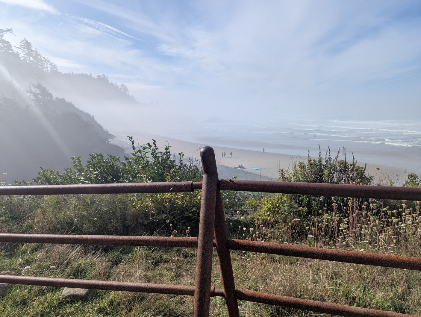 RV rental vacation: Misty coastal beach, people, ocean waves, and forested hills seen through a rusty fence.