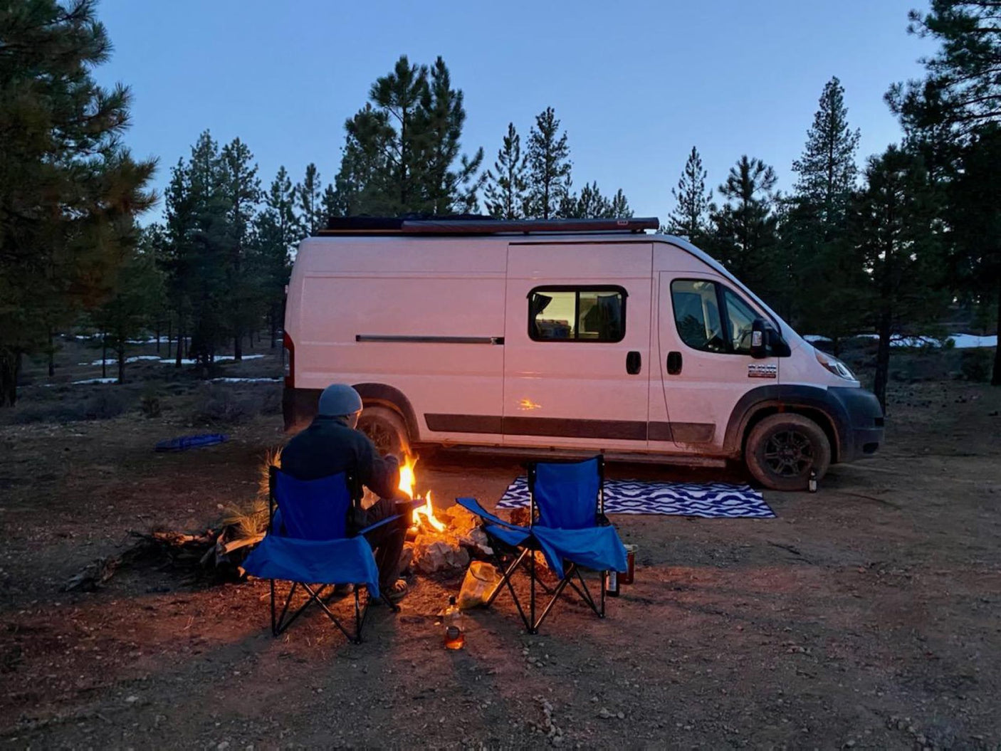 Camping with an RV rental: a person enjoys a campfire next to a white camper van in a wooded area at dusk.