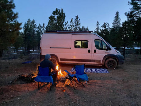 Camping with an RV rental: a person enjoys a campfire next to a white camper van in a wooded area at dusk.