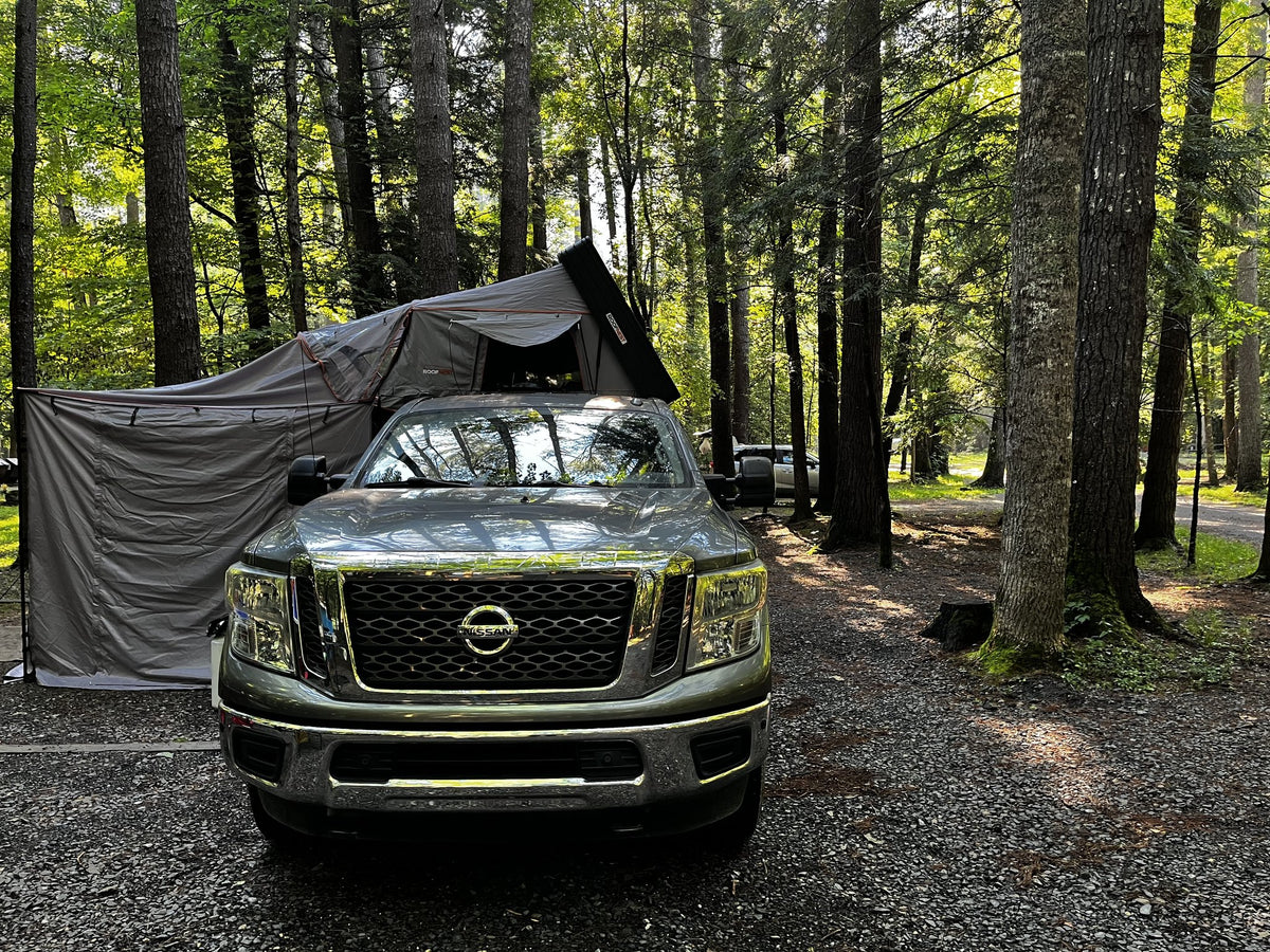 Front view of the rig at a wooded campsite in summer, tent deployed overhead and the awning walls draped down the driver's side to form a shaded side room — photo 5