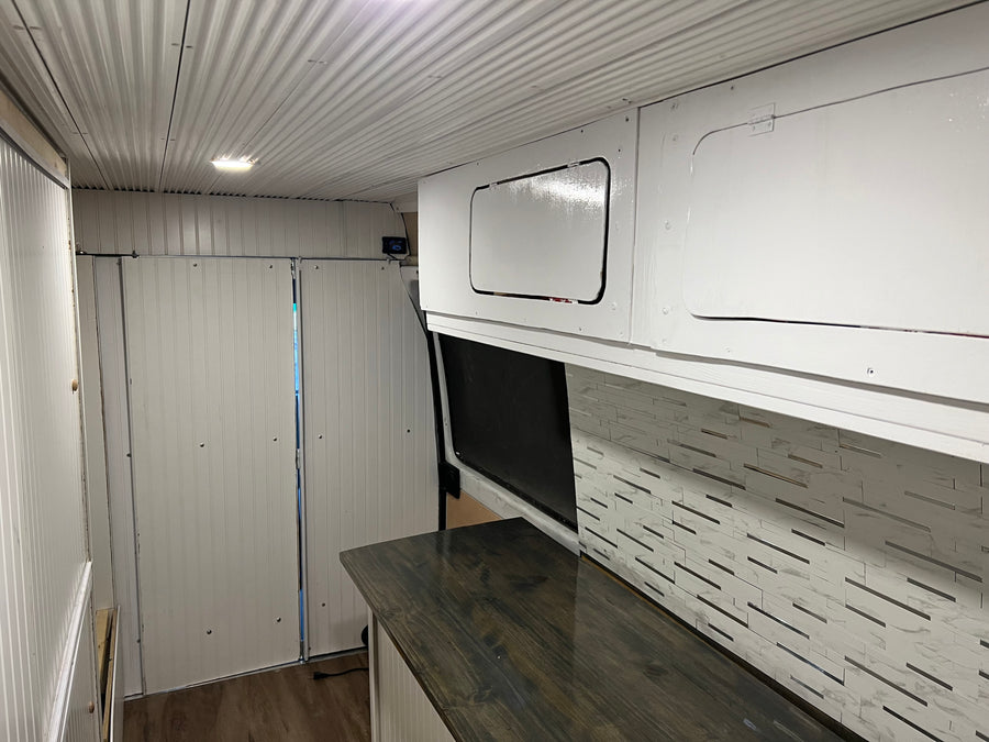 RV rental: Camper van interior featuring white corrugated ceiling, panelled walls, overhead cabinets, dark counter, and tiled backsplash.