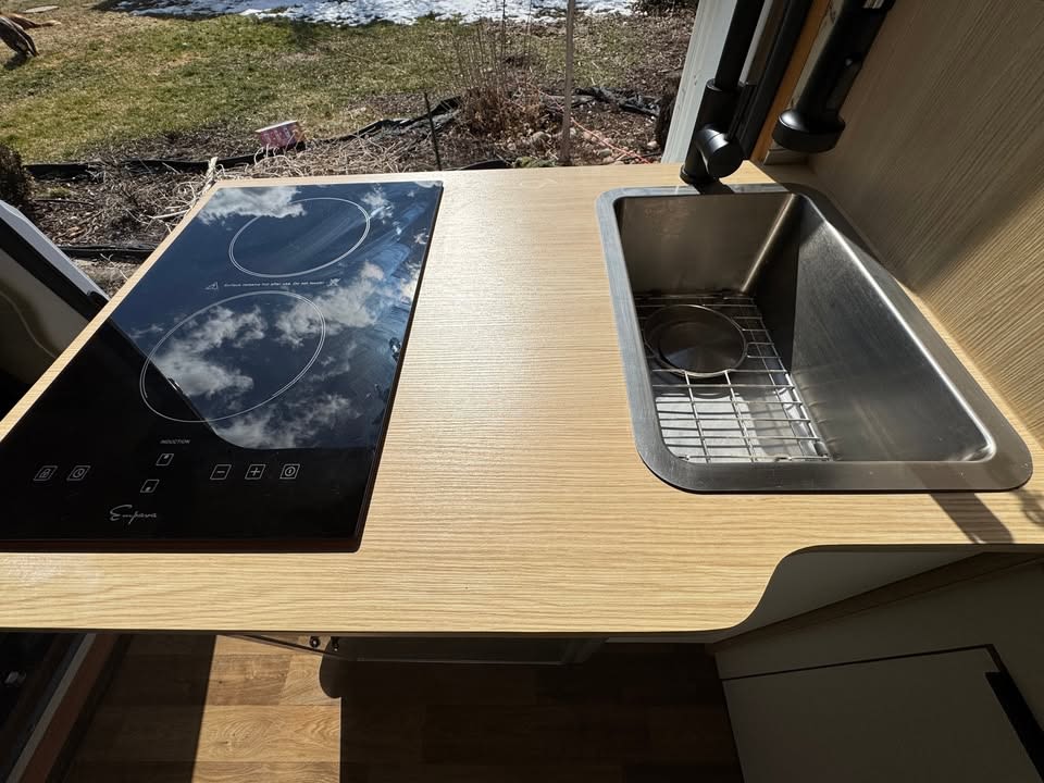 RV rental kitchen features a sleek induction cooktop and stainless steel sink set in a light wood counter.