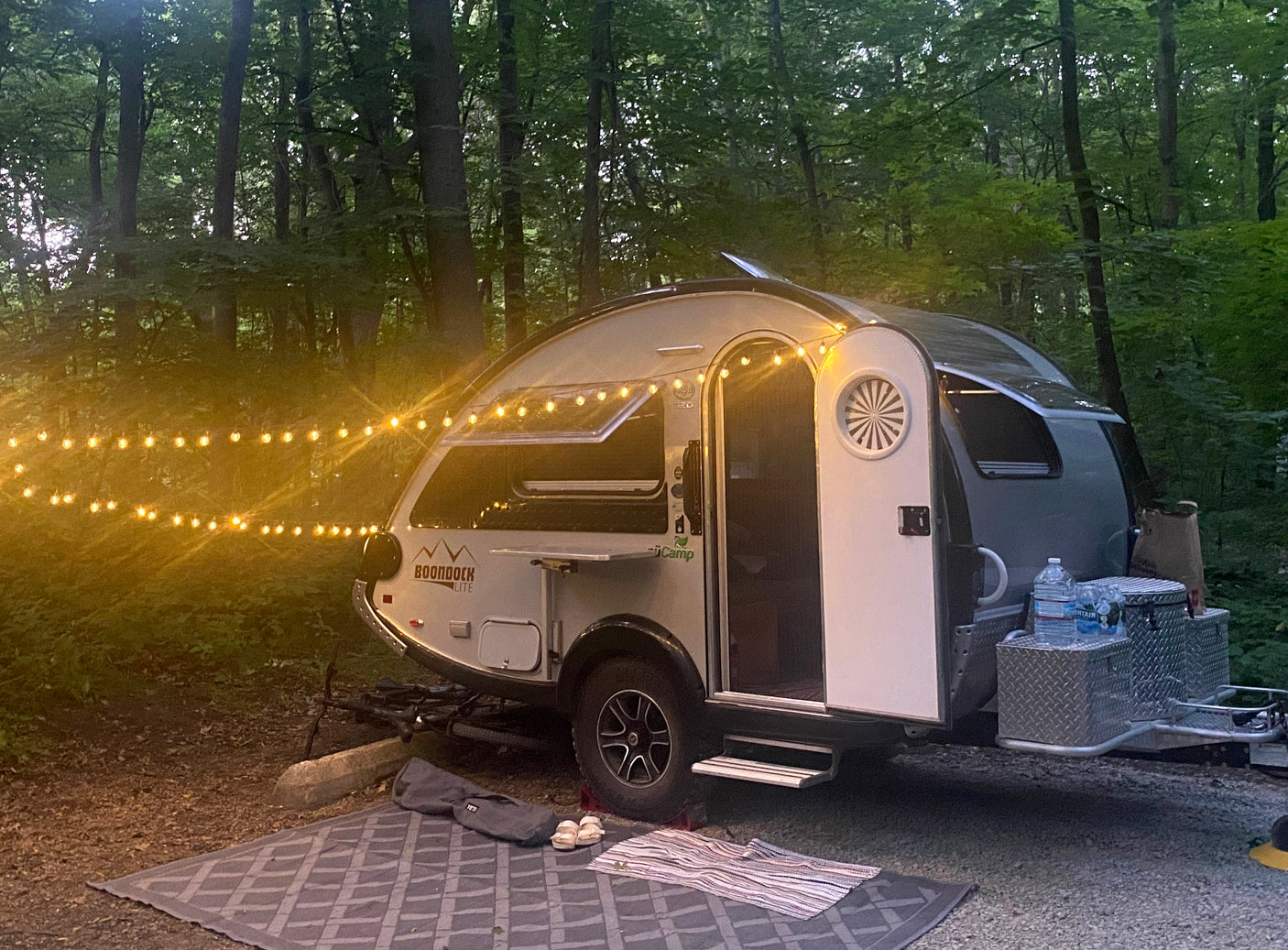 Experience nature! This inviting teardrop RV rental, illuminated by string lights, welcomes you to a peaceful wooded campsite.