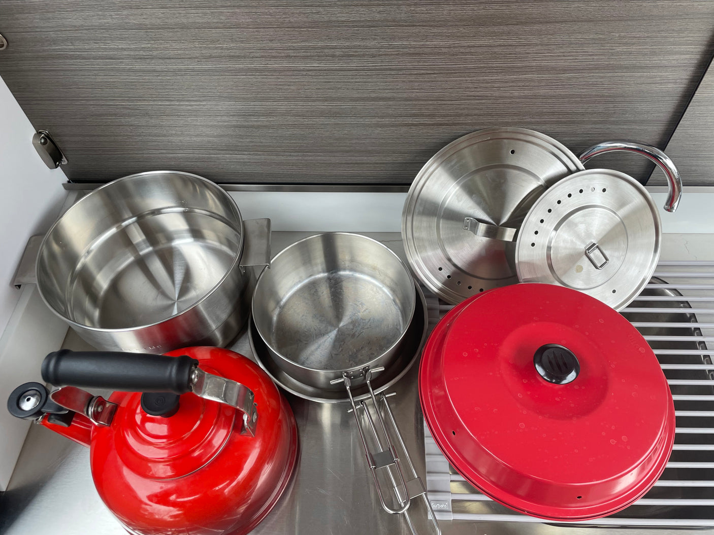 RV rental kitchenware: red kettle, stainless steel pots, and lids on a metal counter, ready for camping meals.
