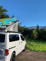 White pop-top RV rental campervan with paddleboards on its roof, parked amidst lush greenery with mountains in the background.