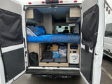 Inside an RV rental: a bed with starry blue sheets, organized under-bed storage, and access to water/power connections.