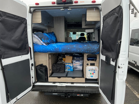 Inside an RV rental: a bed with starry blue sheets, organized under-bed storage, and access to water/power connections.