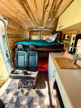 Custom wood paneling, elevated bed, dual swivel seats, and a compact kitchen counter with sink.