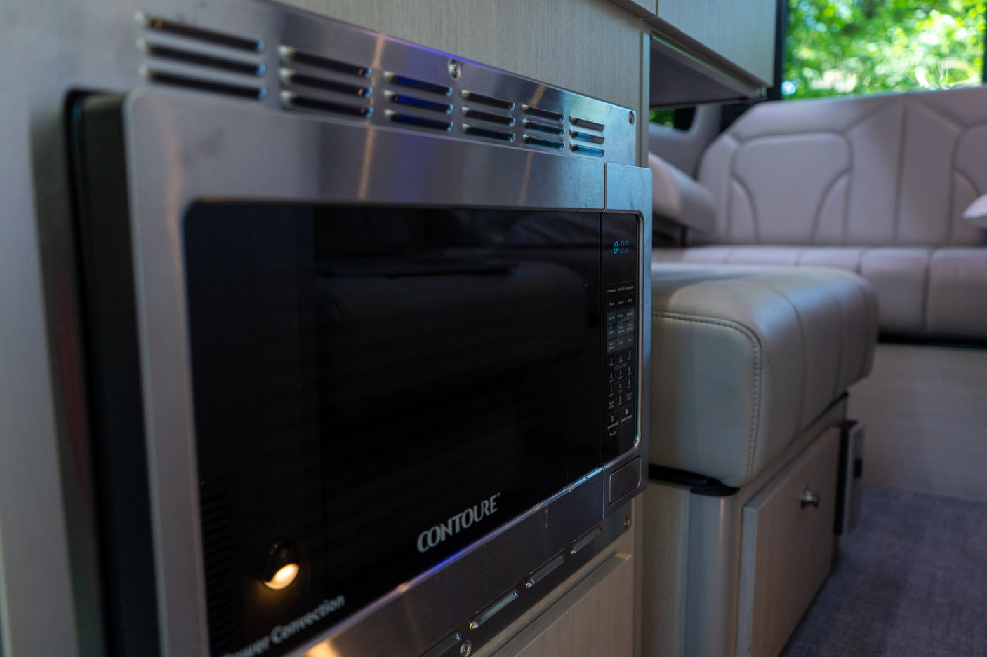 A sleek, stainless steel CONTOURE microwave oven in the modern kitchen area of an rv rental, with plush seating visible in the background.
