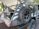 This gray teardrop rv rental features an open outdoor kitchen with sink, stove, microwave, & a spare tire, perfect for camping.