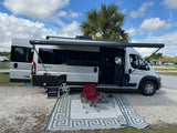 RV rental campervan at a sunny campsite with extended awning. A mat, red chair, and pet bed offer outdoor comfort.