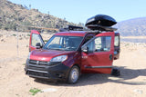 An **rv rental** red Ram ProMaster City van, equipped with an awning & roof box, parked by a lake in a rocky landscape.