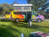 A colorful pop-top RV rental camper van on grass under a blue sky, with a person and camping gear visible.