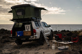 Explore with this RV rental: White Toyota pickup with rooftop tent, ladder, and full camp setup on a rocky beach.