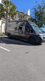Dark gray Ram ProMaster RV rental camper van parked on a sunny day near residential buildings and palm trees.