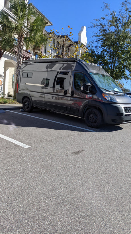 Dark gray Ram ProMaster RV rental camper van parked on a sunny day near residential buildings and palm trees.