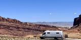 An RV rental, a white camper van with bikes, sits parked in a stunning desert landscape of red rock mountains under a blue sky.