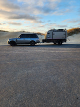 RV rental: A grey SUV tows a rugged off-road camper on an asphalt road by sandy dunes under a cloudy sky.