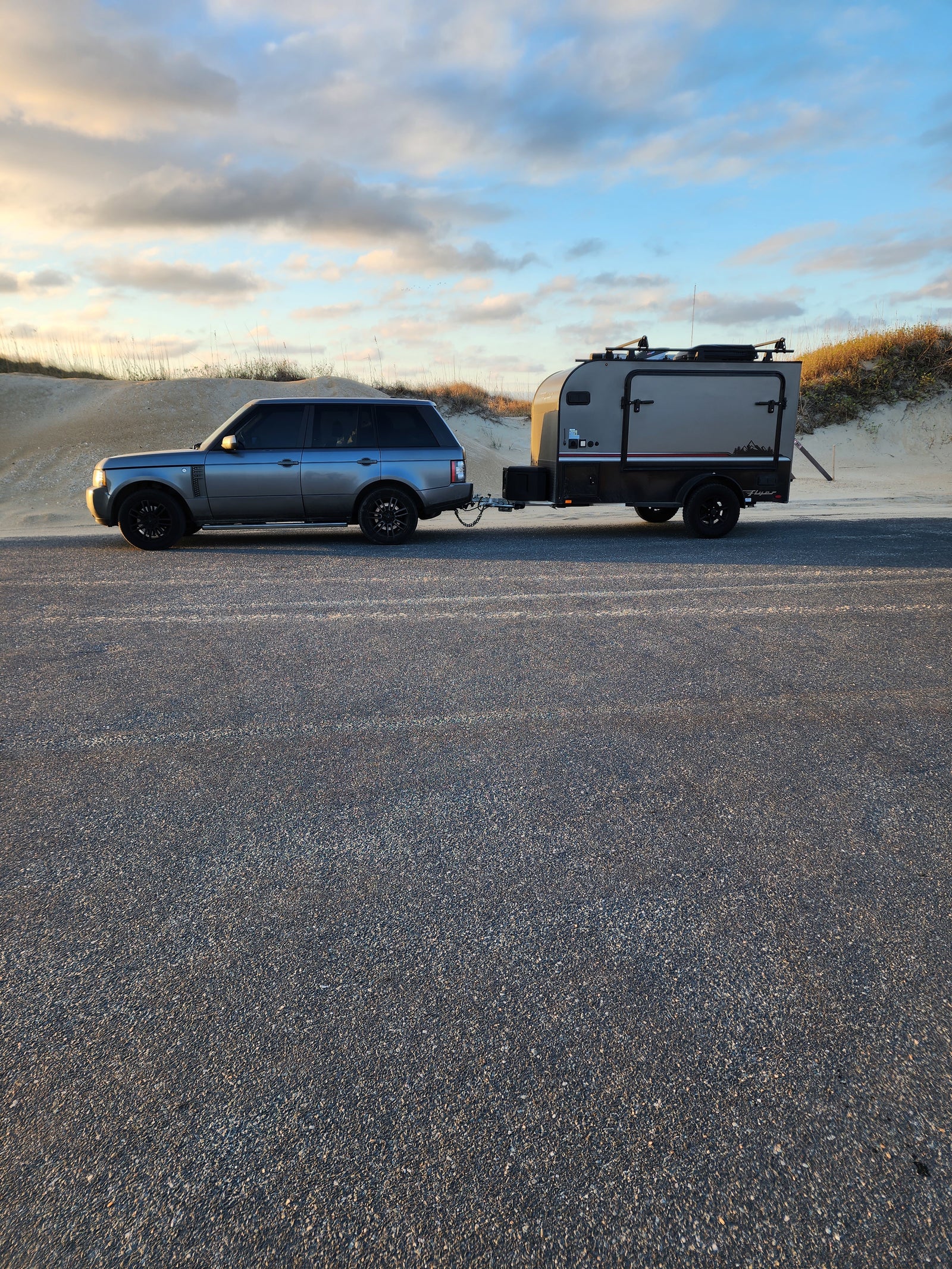 RV rental: A grey SUV tows a rugged off-road camper on an asphalt road by sandy dunes under a cloudy sky.