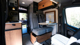 Explore this RV rental: Modern camper van interior featuring swiveling captain's chairs, dining table, kitchenette, and bed.