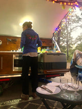 A person cooks in an RV rental's outdoor kitchen at dusk, illuminated by string lights. Dinner prep while camping.