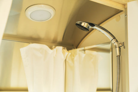 Compact shower in an RV rental, featuring a chrome shower head and ceiling light, with a white shower curtain visible.