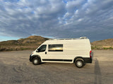 White RV rental van on a gravel lot, reflecting a vibrant sunset under a cloudy sky with hills.