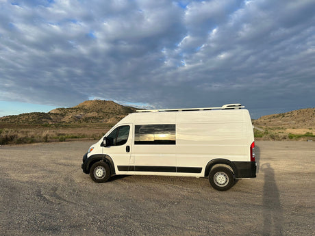 White RV rental van on a gravel lot, reflecting a vibrant sunset under a cloudy sky with hills.