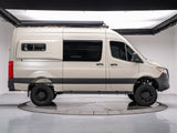 Side profile of a beige off-road camper van with a roof awning, chunky tires, and custom suspension, available for rv rental.