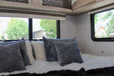 Interior of an RV rental bedroom with soft grey and white pillows on a fluffy bed by large windows.