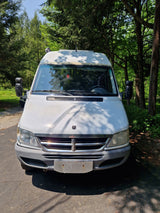 Front view of a white Sprinter camper van, ideal for your next rv rental adventure, parked on a paved lot among trees.