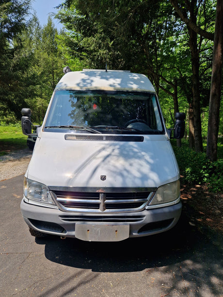 Front view of a white Sprinter camper van, ideal for your next rv rental adventure, parked on a paved lot among trees.