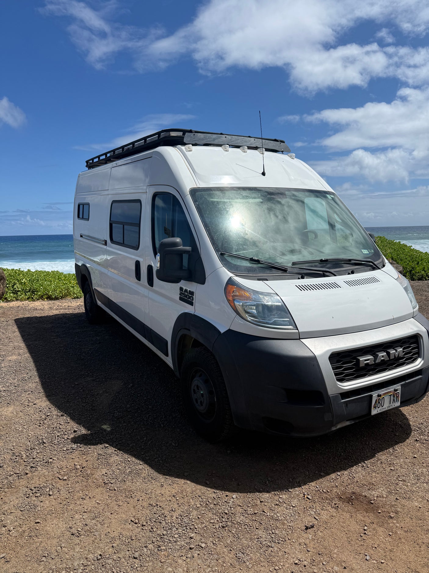 Coastal RV rental: A white Ram camper van with a roof rack, parked on gravel next to a vibrant blue ocean under sunny skies.