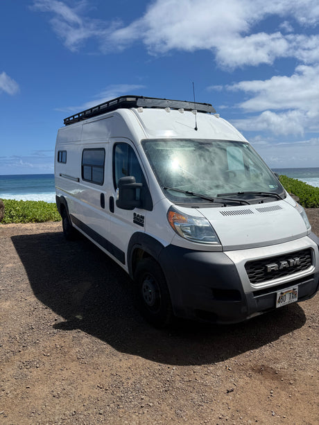 Coastal RV rental: A white Ram camper van with a roof rack, parked on gravel next to a vibrant blue ocean under sunny skies.