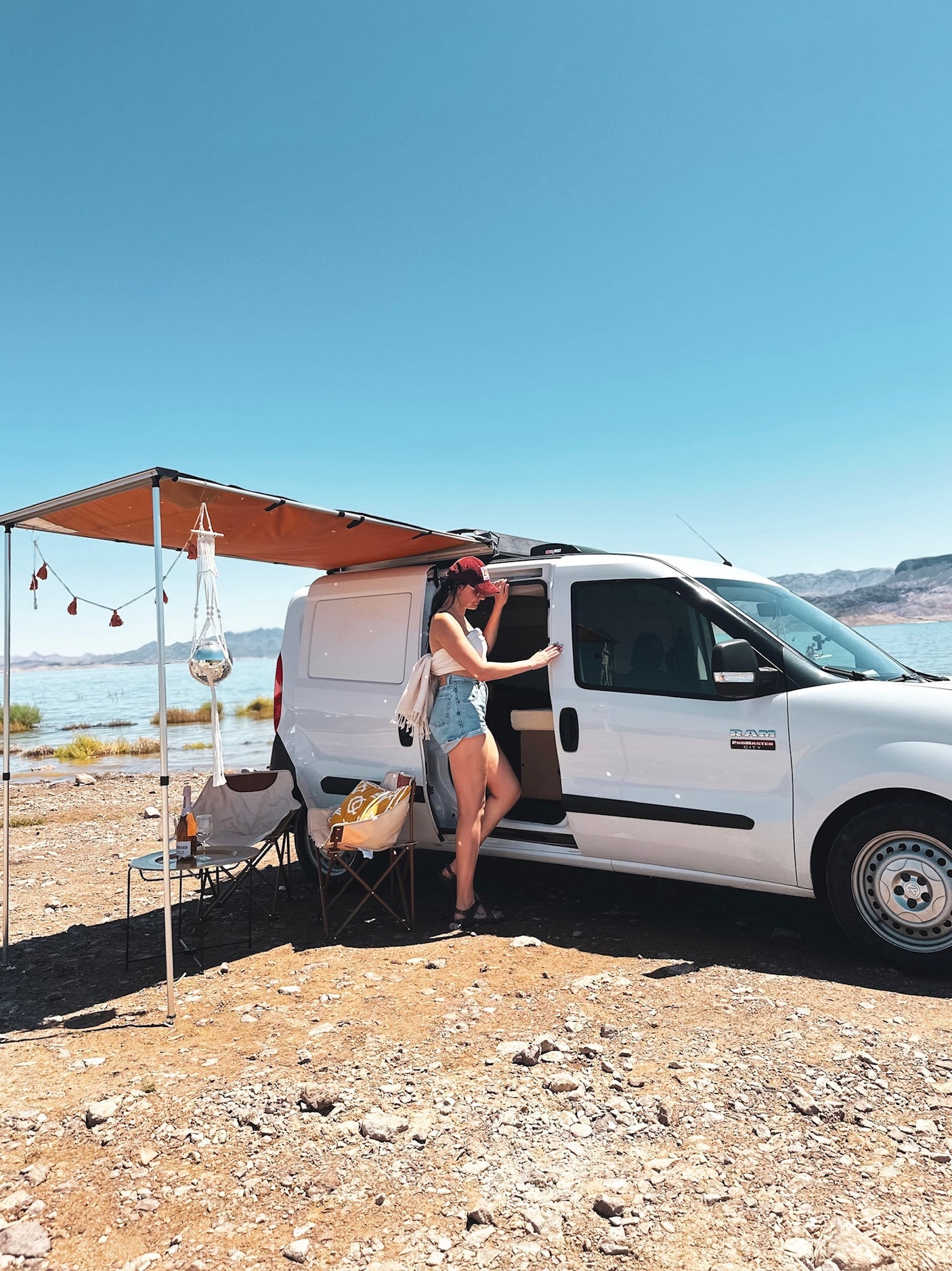 Scenic RV rental adventure: Woman exits white van with awning, camp setup by clear blue lake. Perfect van life!