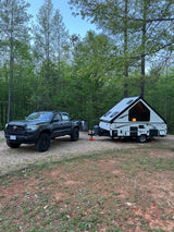 RV rental truck towing an A-frame camper at a wooded campsite, ideal for outdoor adventures.