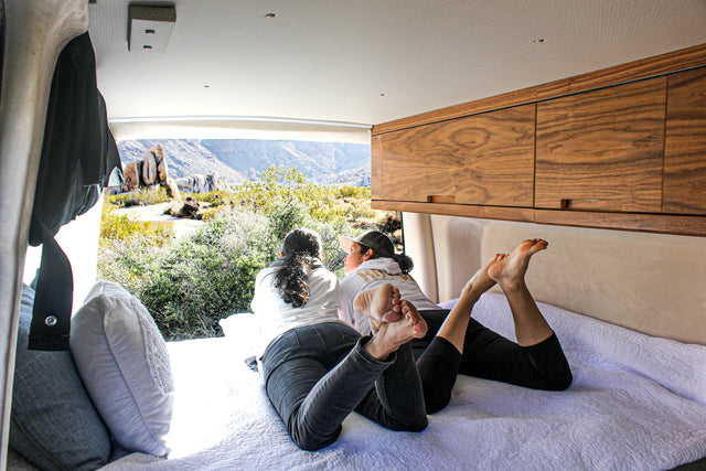 RV rental comfort: Two women relax in a camper van, enjoying desert mountain views from a cozy bed.