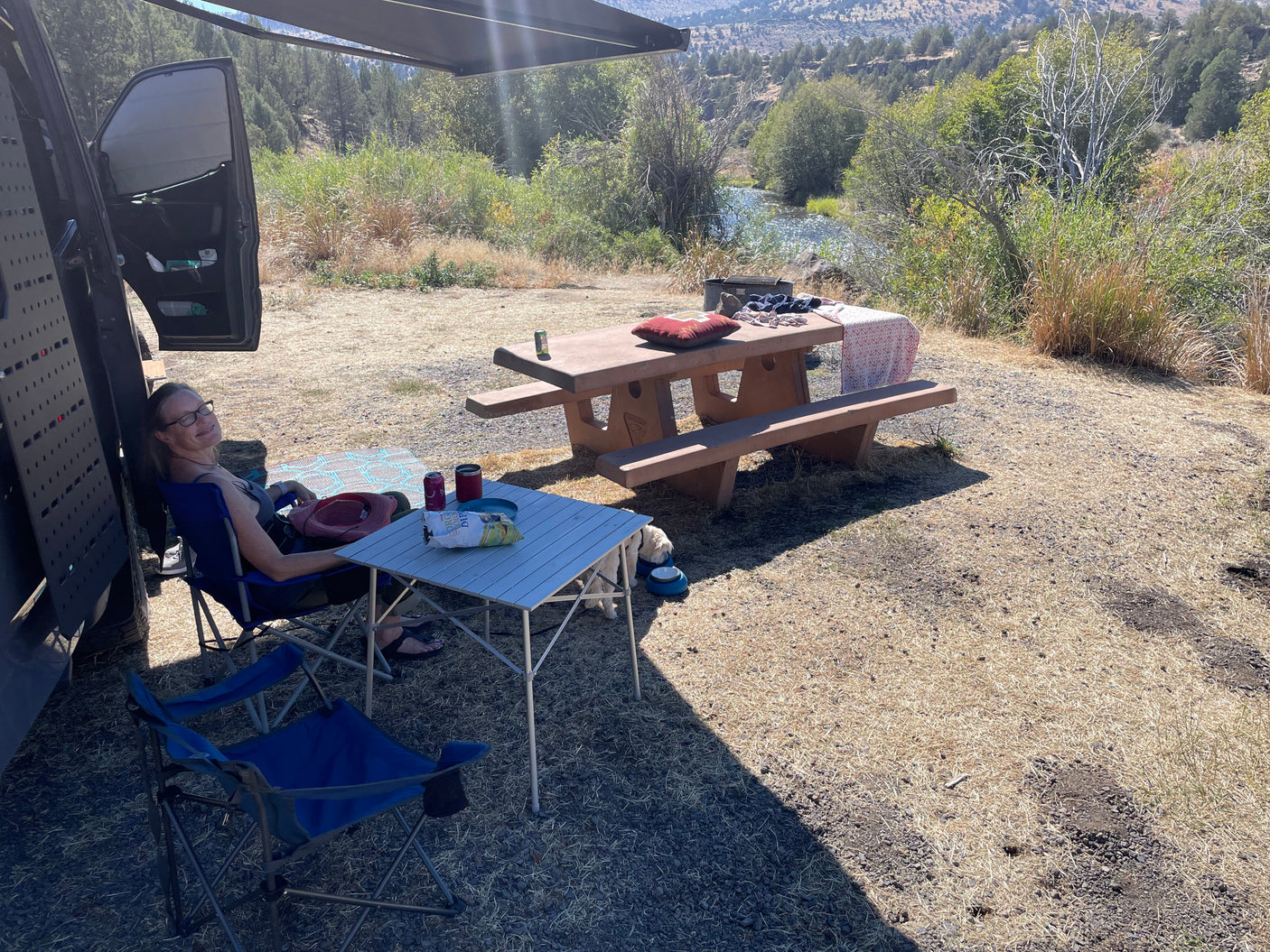 Serene riverside RV rental campsite scene. A woman relaxes by the van, with a dog and picnic table nearby, overlooking the river.