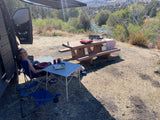 Serene riverside RV rental campsite scene. A woman relaxes by the van, with a dog and picnic table nearby, overlooking the river.