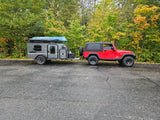 A red Jeep Wrangler tows a gray RV rental trailer with a blue canoe on top, parked in front of a forest displaying autumn colors.
