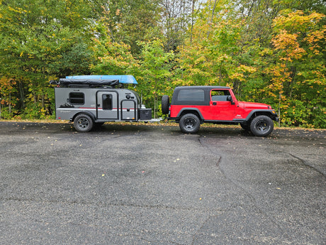 A red Jeep Wrangler tows a gray RV rental trailer with a blue canoe on top, parked in front of a forest displaying autumn colors.