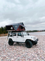 Explore with this adventurous RV rental: a white Jeep Wrangler with a deployed rooftop tent on a rocky shore.