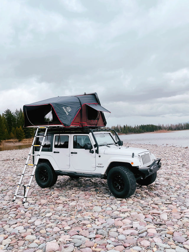 Explore with this adventurous RV rental: a white Jeep Wrangler with a deployed rooftop tent on a rocky shore.
