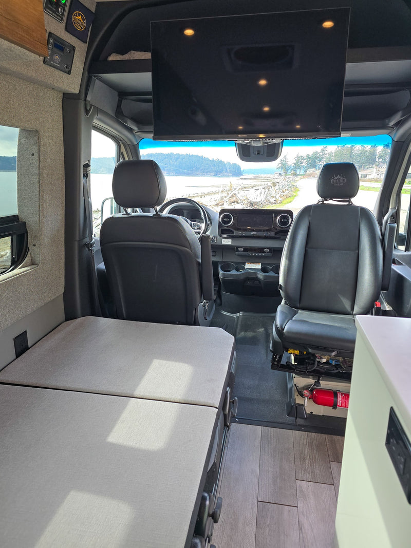 Interior of a clean RV rental, showing the driver's cabin, overhead TV, and flexible living area with a bed. Coastal view outside. — photo 6